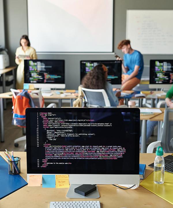 Imagen vertical de una computadora con códigos en el monitor de pie en el escritorio en el aula con estudiantes y maestros en el fondo
